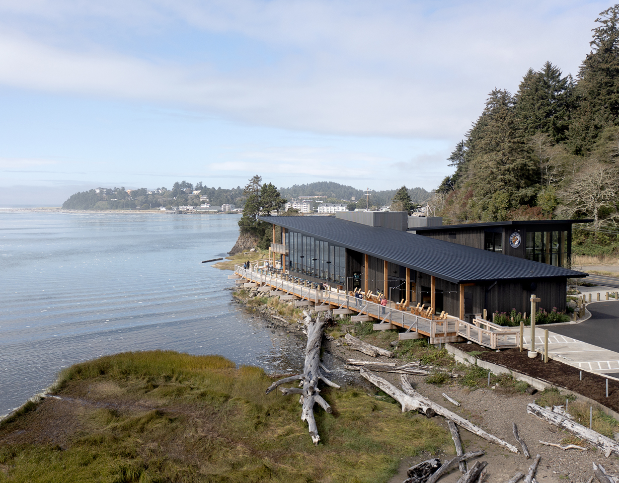 Aerial photograph of a brew pub situated next to a bay on the Oregon Coast. The sky is partly cloudy and large pieces of driftwood lie in the foreground. 